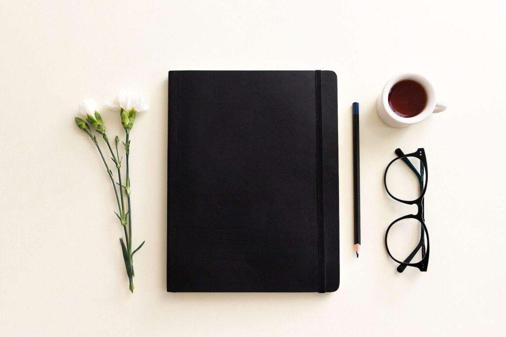 Caderno fechado na mesa com óculos, café, caneta e flor